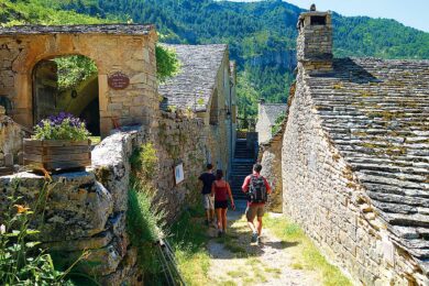 Drei Personen wandern in durch ein kleines Dorf in Gorges Du Tarn. Im Hintergrund sind Berge zu sehen